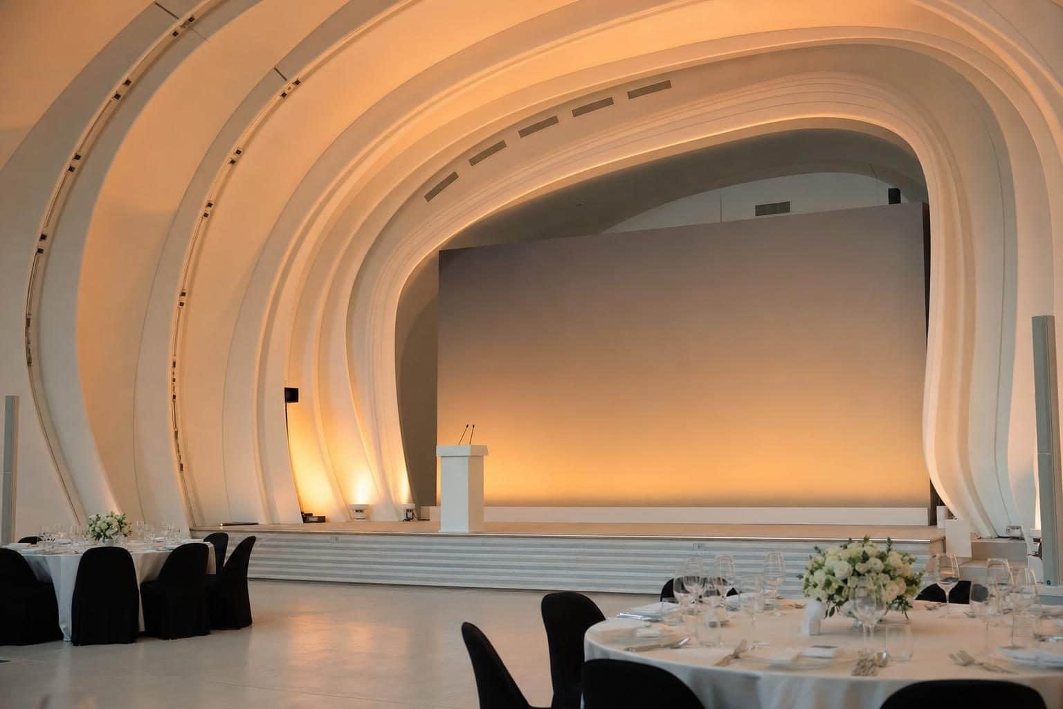 Clean modern stage with blank lectern, soft amber gradient backdrop, framed by Zaha Hadid parametric curves sweeping overhead, Heydar Aliyev Center