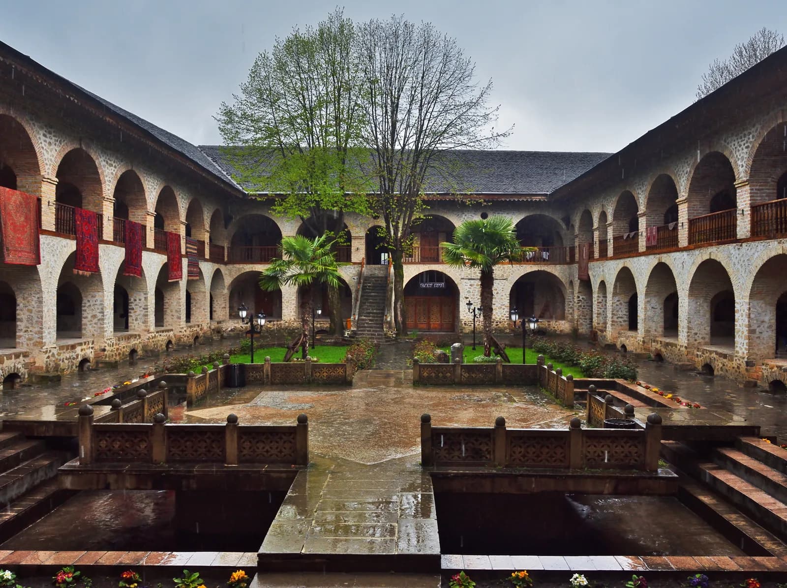 The arcaded inner courtyard of the Sheki Caravanserai after rain, carpets hung from the upper gallery