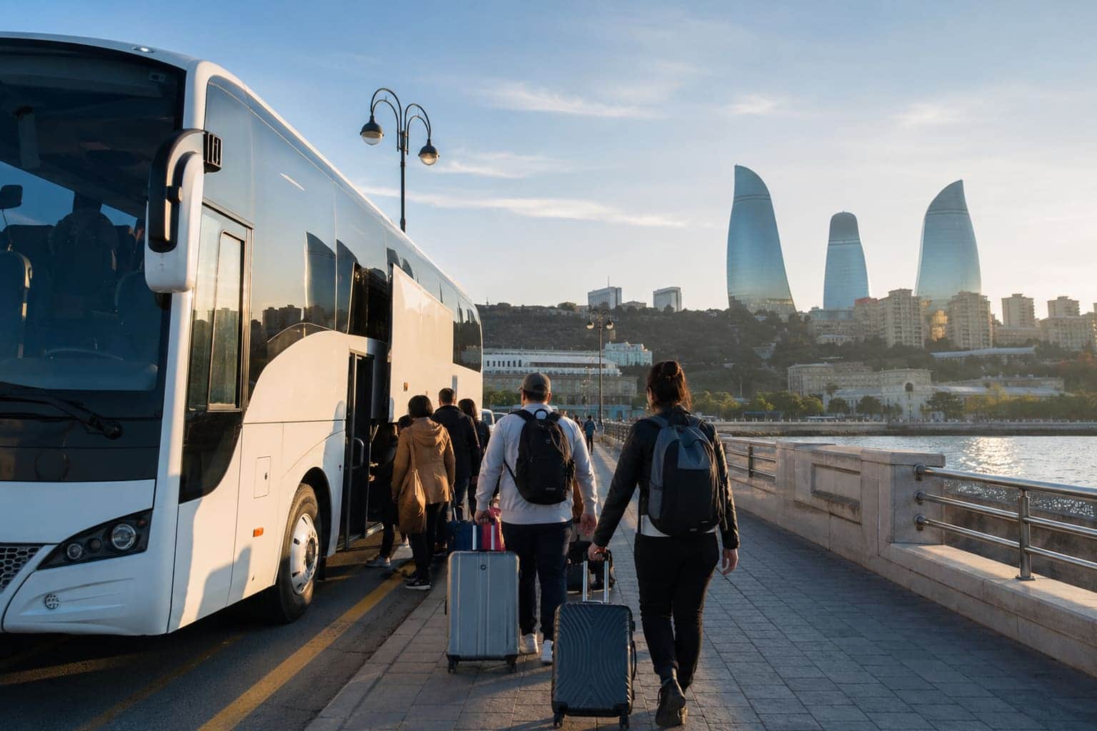 Travelers with suitcases boarding a white tour coach on Baku's seaside boulevard, with the Flame Towers in the background