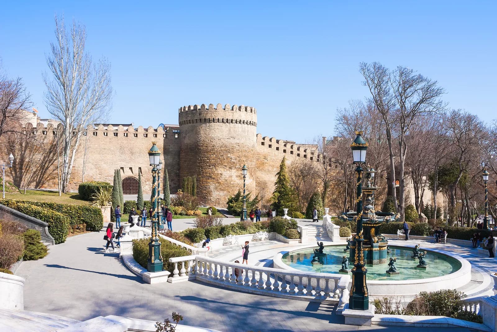 Visitors walking below the fortress walls of Icherisheher, the Old City of Baku, beside an ornamental fountain in the garden